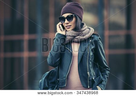 Young fashion woman in black leather jacket talking cell phone  Stylish female model in sunglasses beanie and snood scarf on city street