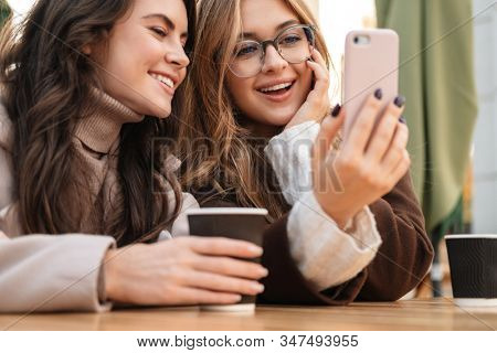Two cheerful attractive women friends sitting at the cafe outdoors, looking at mobile phone while drinking coffee