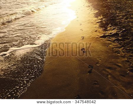 Human Footprints On The Sand Of A Beach On The Shore With Foamy Waves And The Light Of The Setting S