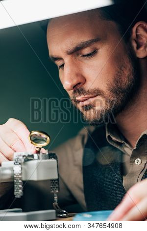 Selective Focus Of Handsome Watchmaker Working With Magnifying Glass And Wristwatch On Movement Hold