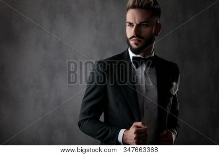 sexy businessman wearing black tuxedo standing and fixing his jacket while looking  away with a dramatic look on his face on gray studio background