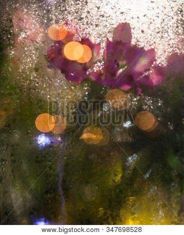 Flowers And Light Garlands Behind The Wet Glass. Moist Window. Water Drops On The Glass. Blurred Lig