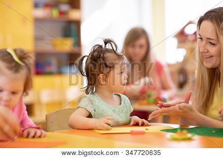 Worker With Babies In Creche. Kids Group On Lesson In Kindergarten