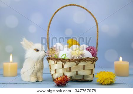 The Easter Bunny Sits Next To A Basket Full Of Eggs, Decorated With Dried Flowers. Burning Candles I