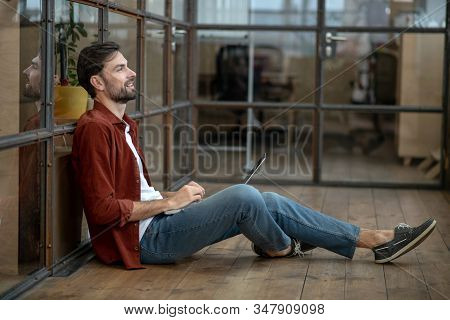 Beraded Handsome Man In A Brown Shirt Sitting On The Floor And Looking Thoughtful