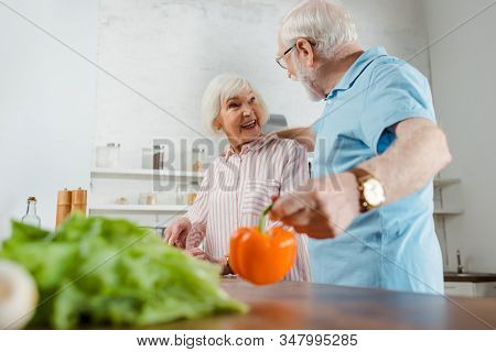 Selectie Focus Of Senior Man Hugging Smiling Wife While Holding Bell Pepper By Kitchen Table