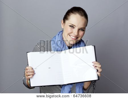 Portrait of a smiling young woman, with long brunette hair, on gray studio background, holding an open photobook or album in her hands 