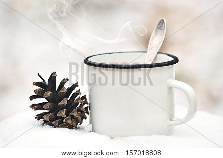 Old mug with a hot drink and with a fir-cone on a wooden stump in the snow in the outdoors. Photo in vintage color image style. Coffee break outdoors. Morning Still life. Morning at the campsite.