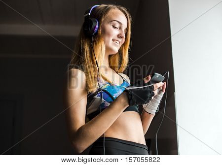 Attractive blonde girl in purple headphones with bottle in hand listening musci near the window
