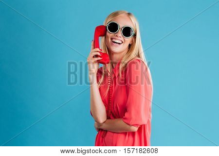 Happy stylish blonde woman talking on red phone and looking at camera isolated on a blue background