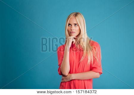 Portrait of a pensive thougtful pretty blonde girl in red dress thinking about something isolated on a blue background