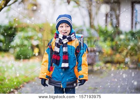 Little school kid boy of elementary class walking to school during snowfall. Happy child having fun and playing with first snow. Student with backpack in colorful winter clothes catching snowflakes.