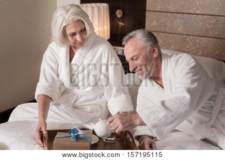 Having breakfast together. Cheerful smiling aged couple lying on the bed and having a breakfast while expressing joy
