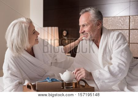 We drinking tea. Cheerful smiling aged couple lying on the bed and having a breakfast while expressing love and care
