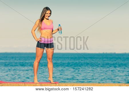 Woman in sportswear takes a break to rehydrate drinking water from plastic bottle resting after sport workout outdoor by seaside
