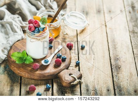 Glass jar of white yogurt with fresh berries, peach and honey on serving board over rustic wooden background, copy space, selective focus, horizontal composition
