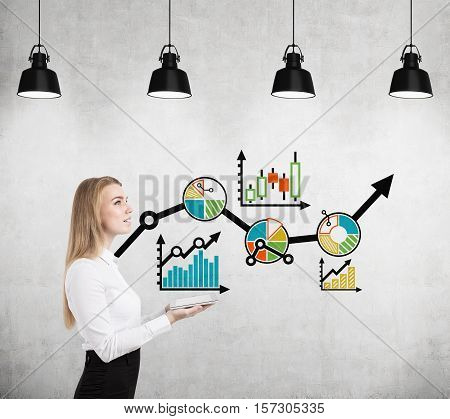 Side view of a woman holding tablet with both hands and standing near a concrete wall with colorful graphs. Concept of growth