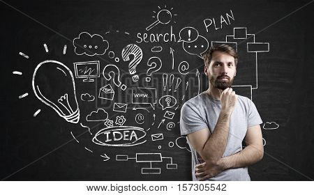 Pensive businessman with beard is standing near blackboard with startup idea sketch on it. Concept of small business planning