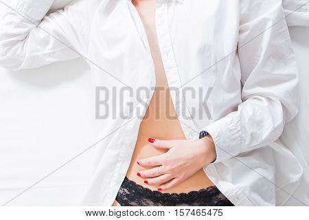 Sexy girl in black lace panties and white shirt lying on bed, hands on belly, body detail, anonymous