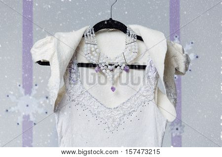 Beautiful white long felted wedding dress with purple details. Warm wool bridal gown with cape and necklace hanging on black hanger. Snow flakes background.