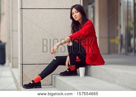 Confident asian businesswoman smiling in the city. Serious beautiful mixed race Asian Caucasian young woman in casual red suite outdoor sitting on steps of building with coffee cup. Next generation