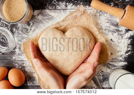 Cooking with love. Female hands holding dough in heart shape top view. Baking ingredients on the dark wooden table