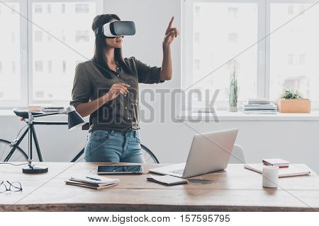 Innovation in business. Confident young woman in virtual reality headset pointing in the air while standing near her working place in office