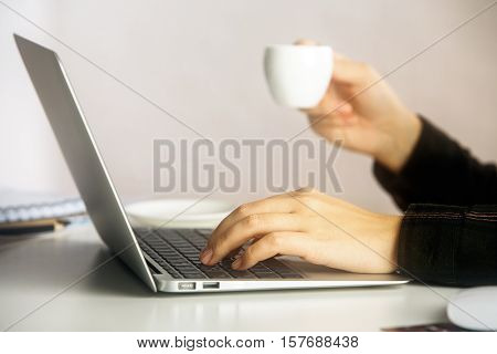 Side view of girl's hands holding coffee cup and typing on laptop keyboard placed on white office desktop. Communication concept