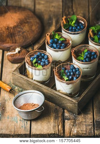 Homemade Tiramisu dessert in glasses with cinnamon, mint and fresh blueberries in wooden tray and sieve with cocoa powder over rustic wooden background, selective focus, vertical composition