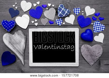 Chalkboard With German Text Valentinstag Mean Valentines Day. Many Blue Textile Hearts. Grey Wooden Background With Vintage, Rustic Or Retro Style. Black And White Style With Colored Hot Spots