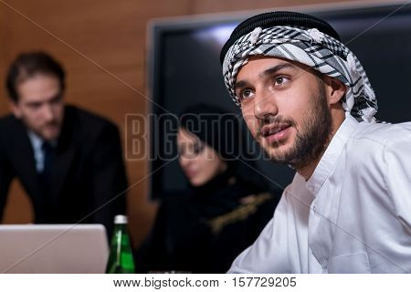Dreams come true. Positive delighted attractive man wearing the white robe and keffiyeh on his head and looking aside while his colleagues discussing project in the backgorund
