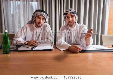 Felling happiness. Two positive delighted attractive men in white robe and keffiyeh on the head sitting at the table discussing and noting some points in the conference hall