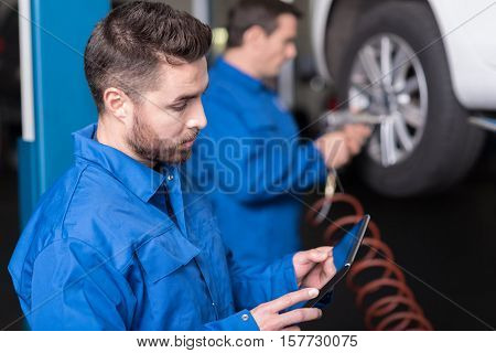 Find out the solution. Handsome confused yong car mechanic using tablet while his colleague changing car wheel in the background