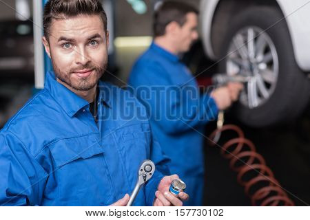 Best equipment in use. Pleasant professional car mechanic holding tools and looking straight while his colleague reparing car in the background