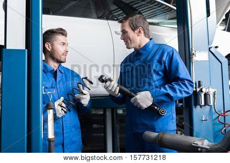Discuss it. Pleasant professional mechanics holding tools and spring while reparing car in auto service