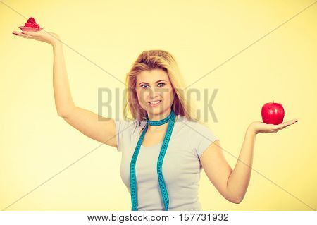 Restrictive diet temptation losing weight concept. Woman choosing between sweet strawberry cupcake and apple having measure tape around her neck. Studio shot on yellow background
