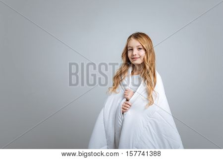 Happ kid. POsitive nice little girl covering with blanket and smiling while standing isolated on grey background