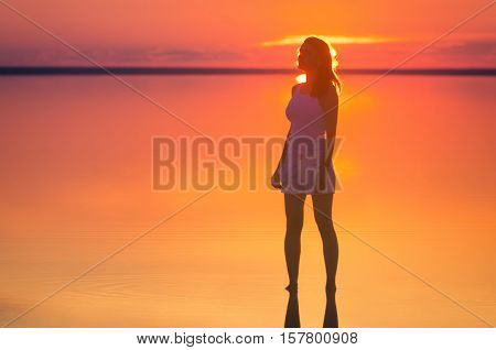 Beautiful female model stands in front of the sun under sunset at seaside. Calm water of salt lake Elton reflects silhouette of woman. Sun goes under horizon. Girl stands alone in water.
