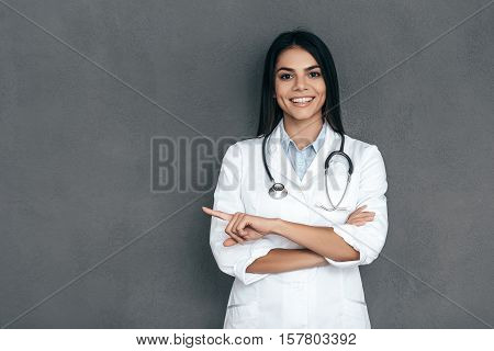 Doctor pointing copy space. Attractive young female doctor in white lab coat keeping arms crossed and smiling while standing against grey background