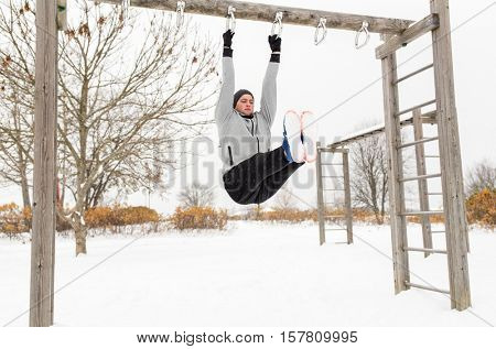 fitness, sport, exercising, training and people concept - young man doing leg pull ups on horizontal bar and flexing abdominal muscles outdoors in winter