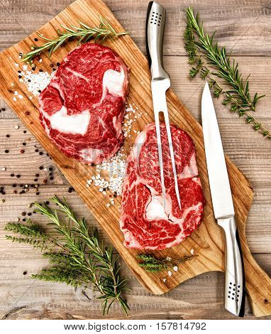 Raw fresh Ribeye Steak with herbs and spices on wooden table. Fork and knife. Food background.