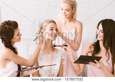 We helping the bride. Happy smiling young bridesmaids sitting in the white bedroom while helping the bride to get ready and expressing joy