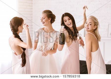 Hen party. Cheerful laughing young girls standing in the white bedroom while having fun and drinking alcohol