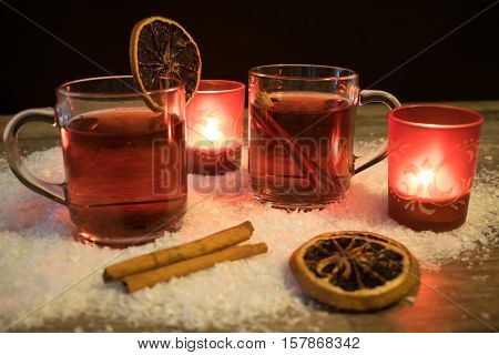 2 glasses with mulled wine in the snow with orange slices cinnamon sticks and 2 red tea light holders with ever a burning tea lights