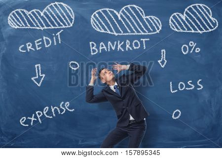 A businessman trying to cover his head with his hands surrounded with clouds and words credit, loss, bankrot, drawn on the blue chalkboard. Bear losses. Face serious challenges. Business difficulties.