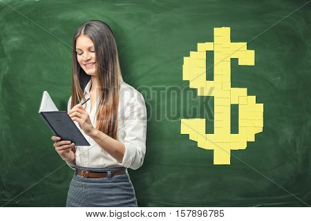 A young businesswoman holding her open day planner and writing something in it and yellow sheets of paper forming big dollar sign on background. Daily planning. Time-management. Secretary work.