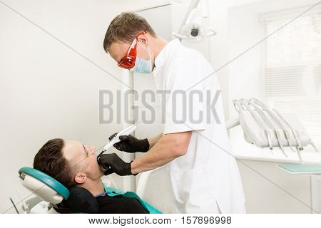 Dentist in the process of work. Male doctor treats a patient tooth using blue; dental; curing; light.