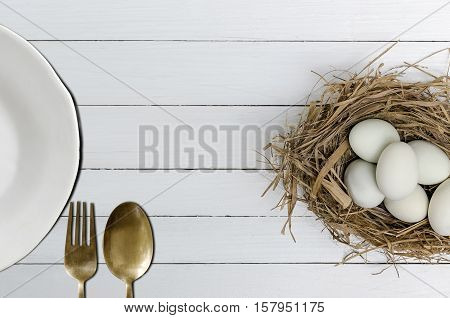 Duck eggs in a grass nest with white plate spoon and fork on white wooden background.