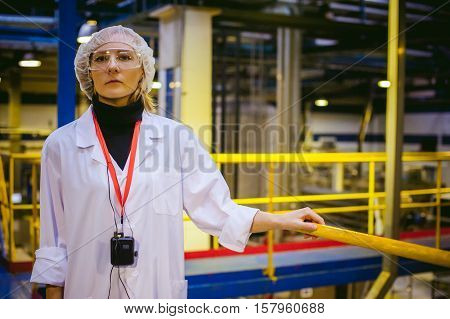 Female Worker On The Production Line Of Beer Factory. Portrait Of A Woman In A White Robe, Standing