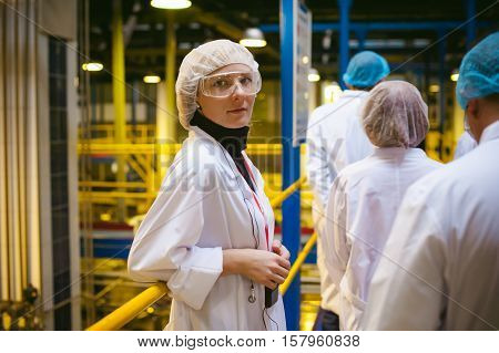 Female Worker On The Production Line Of Beer Factory. Portrait Of A Woman In A White Robe, Standing